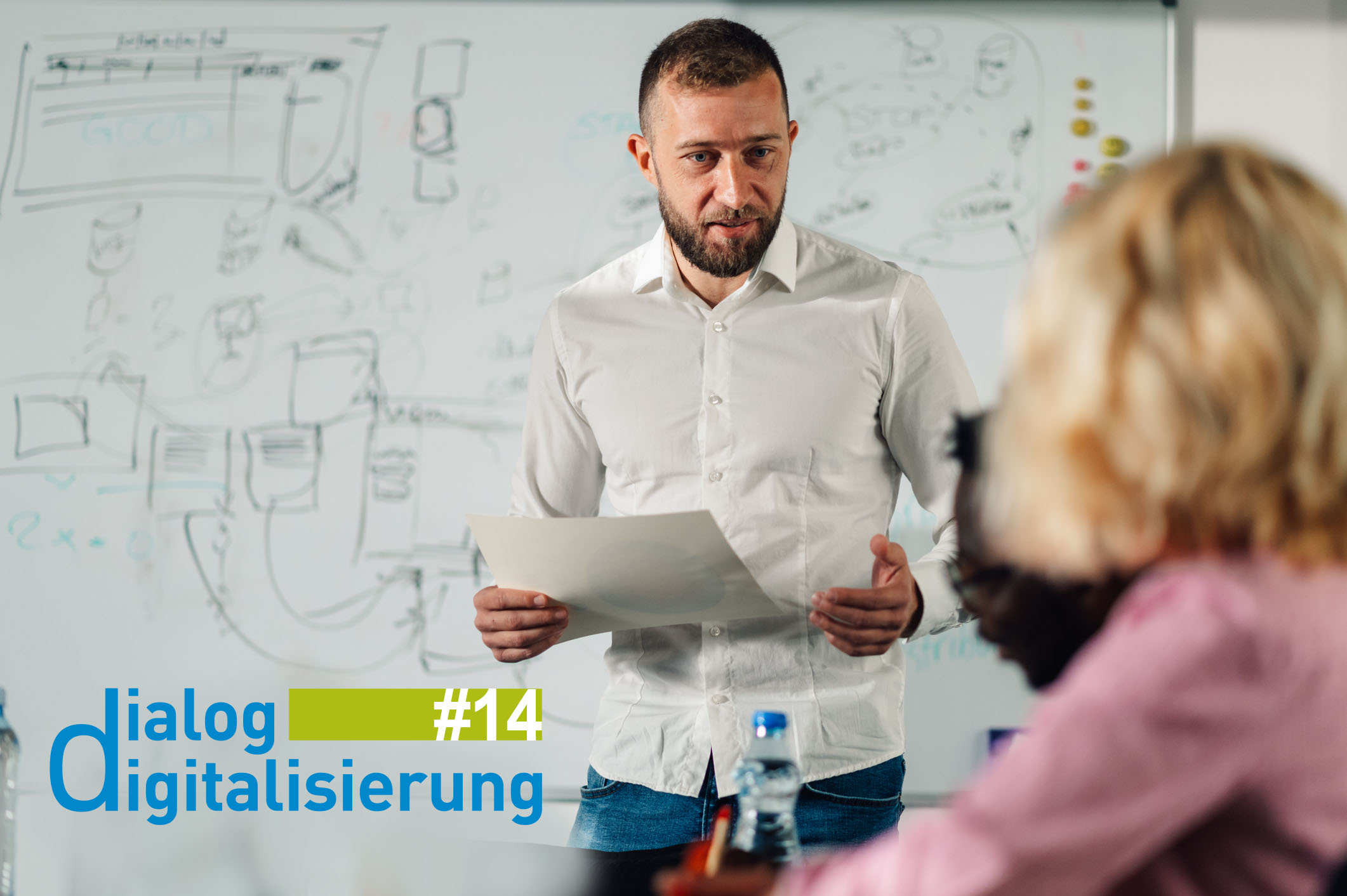 Das Bild zeigt einen Mann mittleren Alters, der vor einem vollgeschriebenen Whiteboard mit Abläufen steht und in Interaktion mit zwei Personen zu sein scheint, die ihm gegenüber sitzen.